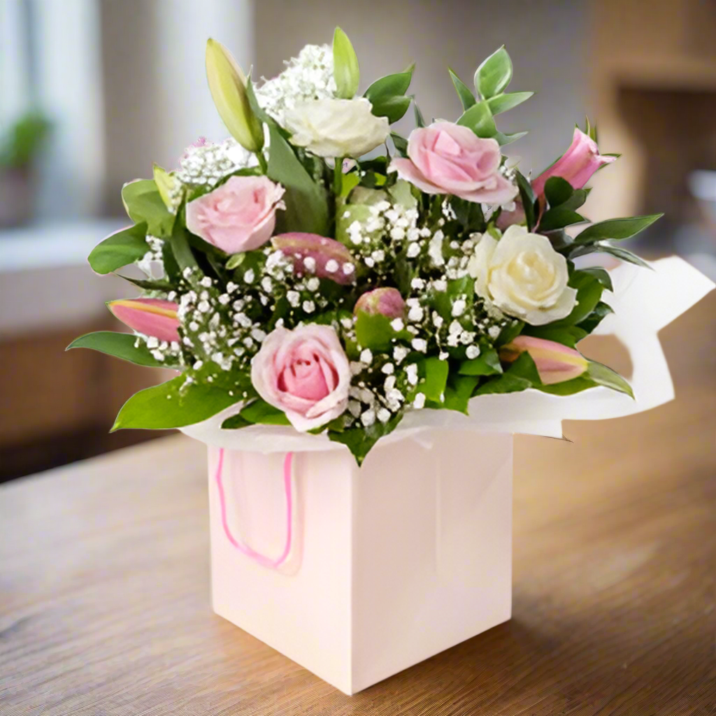 A bouquet of soft pink roses, oriental lilies, and foliage arranged in a white posy bag