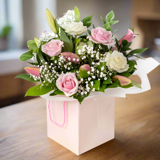 A bouquet of soft pink roses, oriental lilies, and foliage arranged in a white posy bag