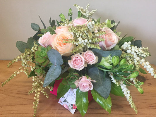 A floral arrangement featuring blush pink roses and lush green foliage in a clear posy box, with water visible at the base