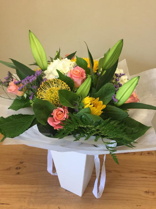 A colorful floral arrangement in a white bag, featuring a variety of flowers including lilies and roses, with green leaves and decorative elements.