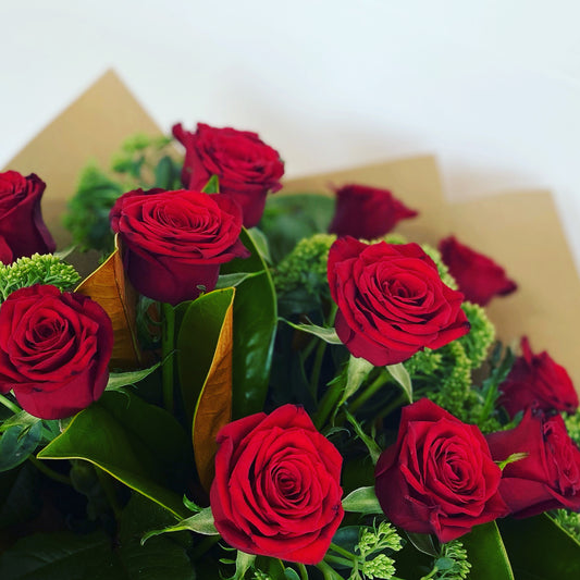 A bouquet of dozen red roses with long stems, arranged with complementary green foliage and packaged in natural brown craft wrap.