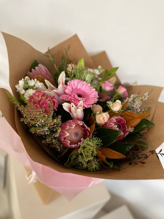 A floral bouquet featuring various pink flowers and green foliage, wrapped in brown paper with pink ribbon.